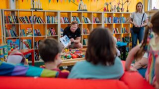 A man reads from a colourful book, surrounded by children