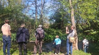 Group of people stood on a river bank looking at the water with trees around them