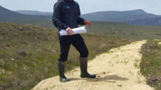 Woman stood in wellies and navy coat on peatland holding paper rolled up in right hand
