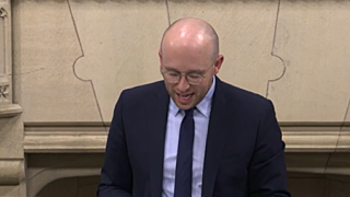 A man looks as his notes, standing up delivering a speech