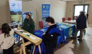 Three people stood looking at an event stand with one person behind the tables talking to them