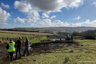 Three people stood with their backs to the camera looking at a muddle channel through a field with a digger in the background