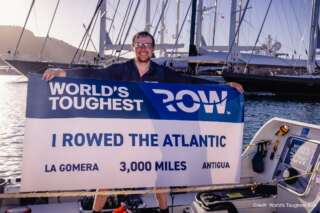 Man stood on stationary rowing boat holding a big sign reading 'I rowed the Atlantic'