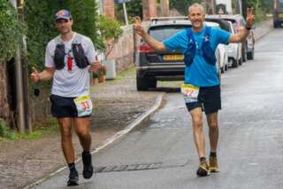 Two white men running along a road towards the camera with the one on the left sticking both thumbs up and the one on the right showing a peace sign on both hands