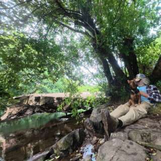 A man leaning against a tree with a dog in his lap by a river