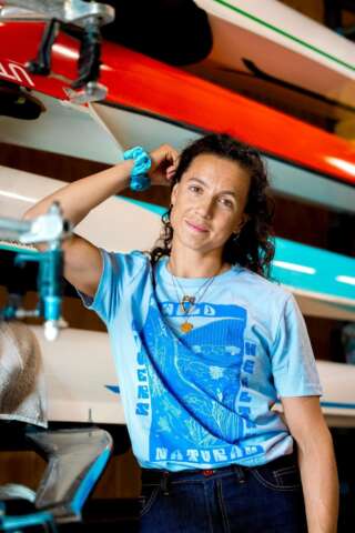 Rower Imogen Grant stands in front of rowing boats, smiling and wearing a light blue teeshirt with a dark blue bold river print