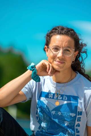 Rower Imogen Grant sits by the water on a sunny day, smiling and wearing a light blue teeshirt with a dark blue bold river print