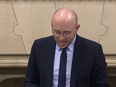 A man looks as his notes, standing up delivering a speech