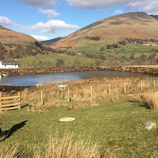 Woman in jeans and black coat stood in field looking at a lake with mountains in background flying a white drone