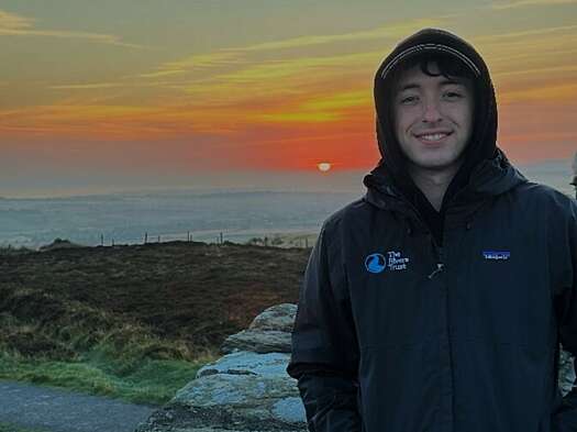 White male in hooded waterproof coat stood with water in background and orange sky
