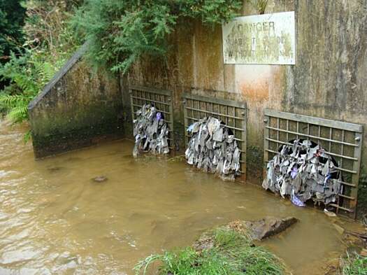 Three sewage pipe ends are open to a river, their grates filled with wet wipes and rubbish