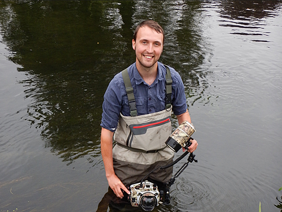 A smiling man stands in a river wearing waders and holding underwater camera equipment