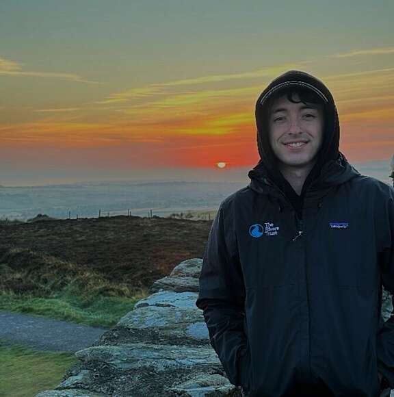 White male in hooded waterproof coat stood with water in background and orange sky