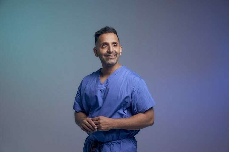 a man smiles off camera, wearing blue medical scrubs