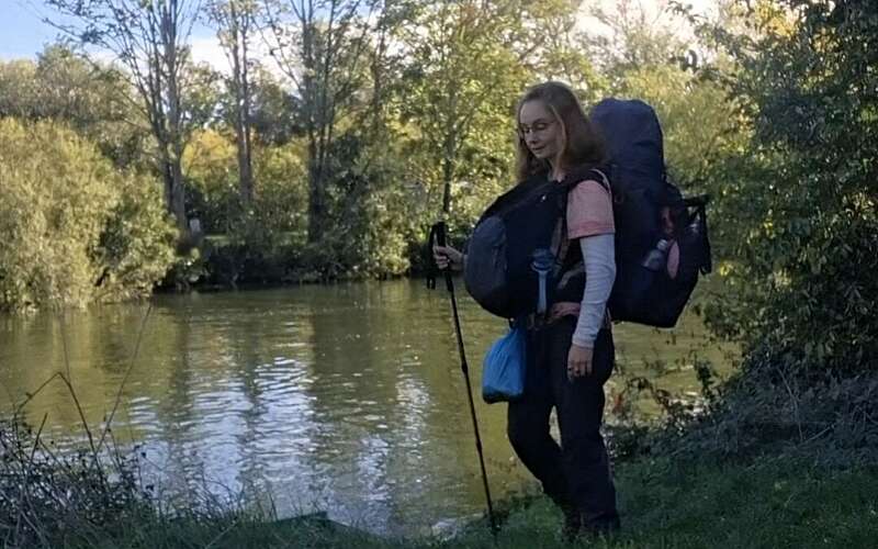 a woman with a backpack and walking pole sands next to a river looking at the water