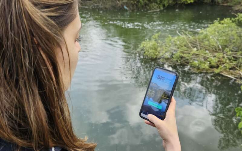 a woman with brown hair holds her phone up with the Big River Watch app on the screen in front of a river