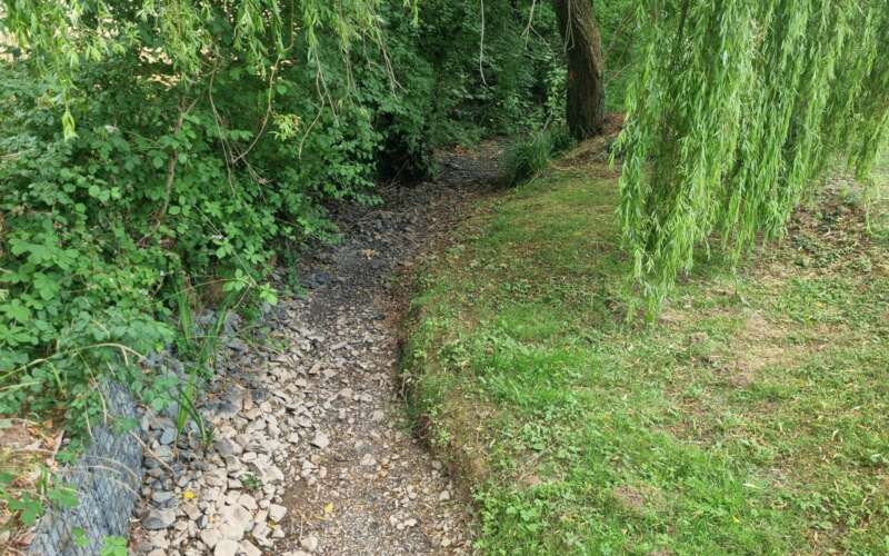 A dry gravel stream bed with grass banks and trees overhanging.