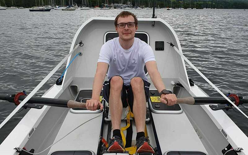 A man in a white teeshirt looks at the camera as he rows a large white boat solo on a lake