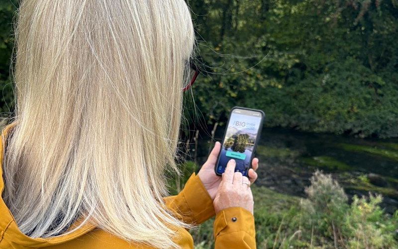 View over woman's shoulder with blonde hair wearing yellow coat holding a phone up in front of a river