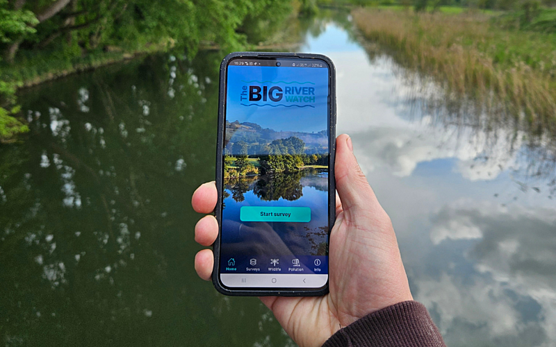 Hand holding a phone in front of a river, displaying the Big River Watch app