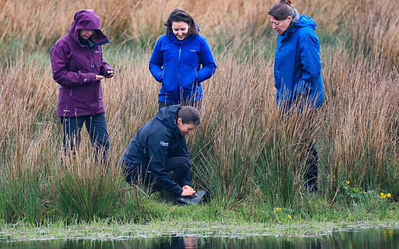 Four women on a river bank one crouched down by the water holding a laptop