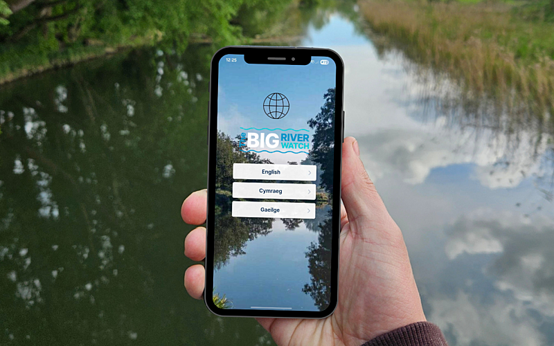 Hand holding mobile phone displaying Big River Watch app with river in background