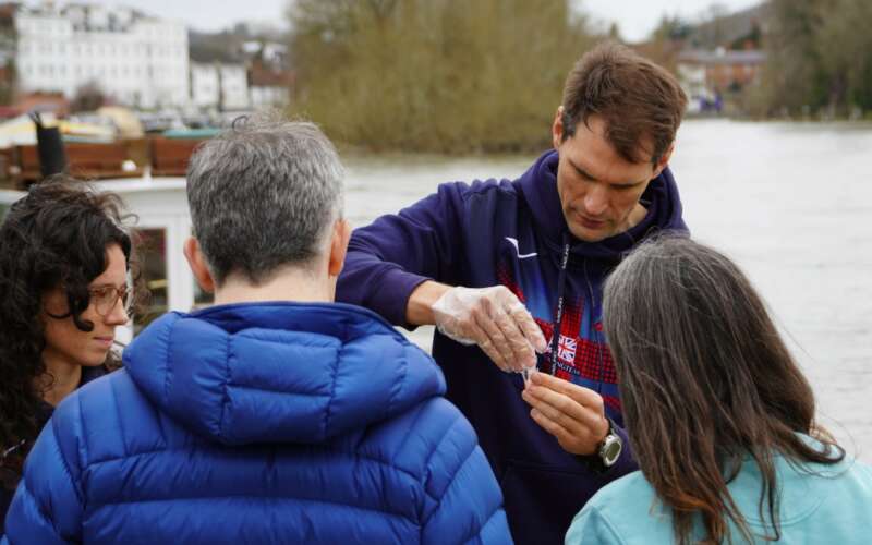 A man with brown hair in a blue hoodie wears plastic gloves while testing a sample of river water for nitrates
