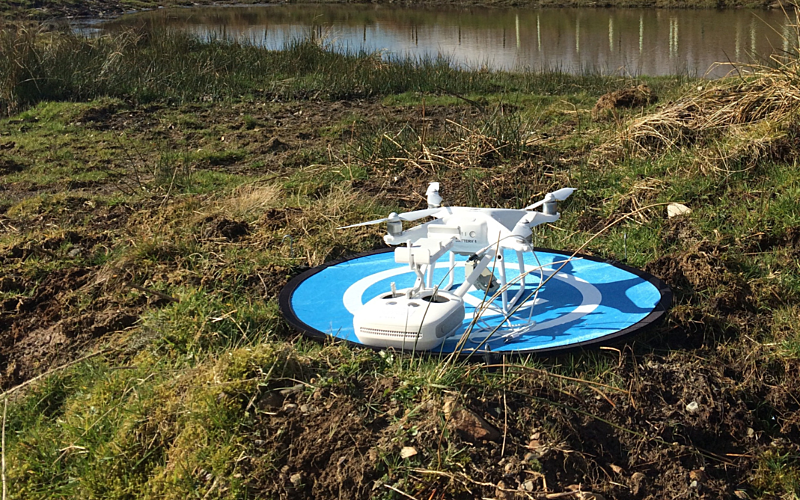 White drone sat on blue landing pad in field with water in background