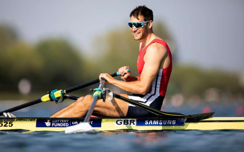 A smiling male rower sits in the single rowing boat, holding his oars in the sunshine