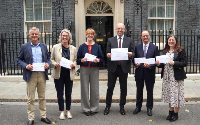 Group of six people, three women and three men stood outside a building with a black door holding envelopes