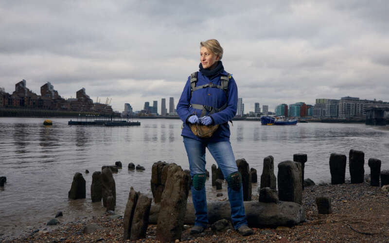 A smiling woman stands on the banks of the Thames, looking into the distance