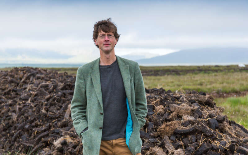 A smiling man looks at the camera, with round glasses and brown hair. In the background is countryside