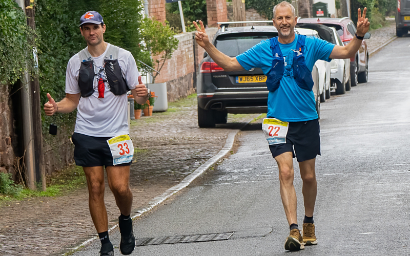 Two male runners, left one in white top with poles, right in blue top, running towards the camera
