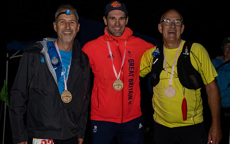 Three male runners stood arm in arm in the dark with medals around their neck