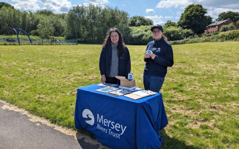 Two women stood behind a table with a cloth draped over it reading Mersey Rivers Trust