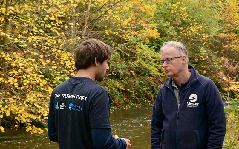Young male with brown hair in navy blue t-shirt on the bank of a river talking to an older male in a dark blue jumper.