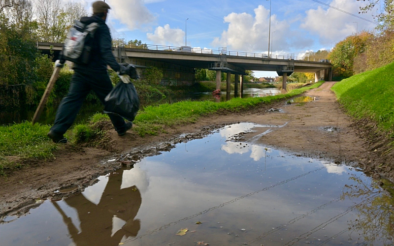 Young male in black coat, wearing a rucksack, walking alongside a river holding a rubbish bag and a litter picker