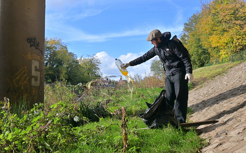Young male in black coat and flat cap on a grassy bank holding up a plastic bottle filled with dirty water