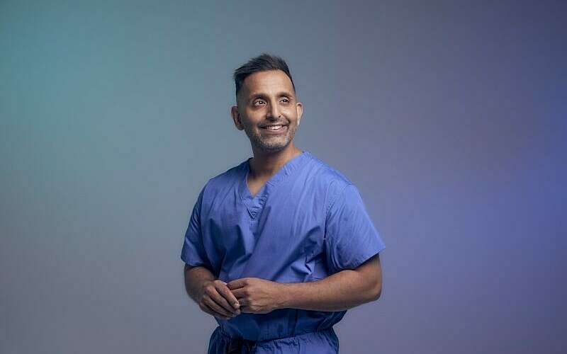 a man smiles off camera, wearing blue medical scrubs