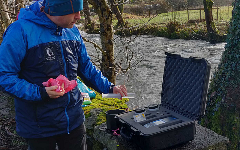 man in blue coat and blue hat stood on banks of river with a case open in front of him containing tubes and testing equipment