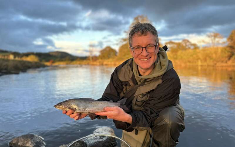 A smiling man holds up a fish by the bank of a river in sunlight