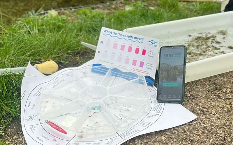a phone displaying the big river watch app, as well as a water testing kit and an invertebrate sampling tray are positioned next to a river