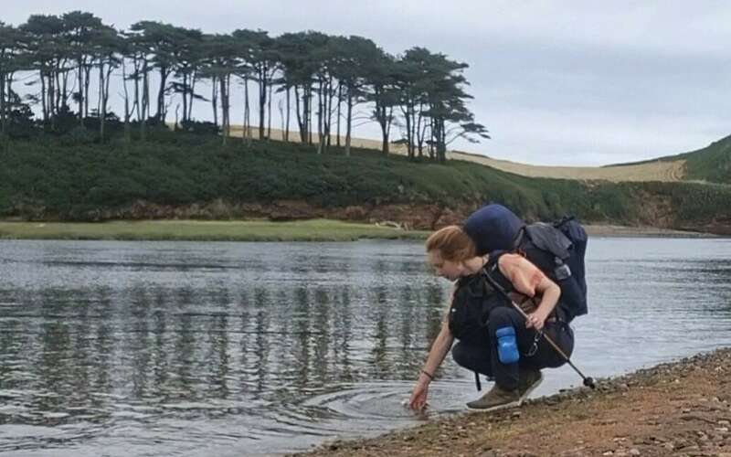 A woman with a backpack bends down to dip her hand in a low river