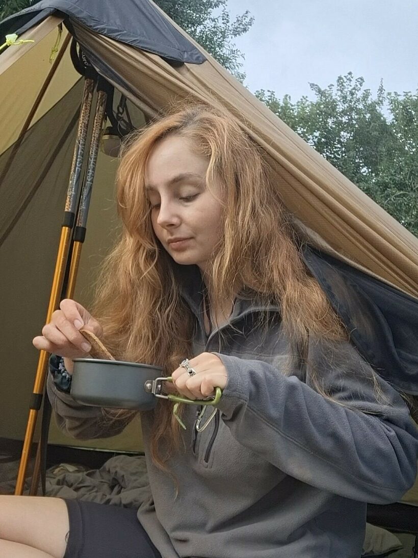 sat at the entrance of a tent, a woman eats from a camping pot