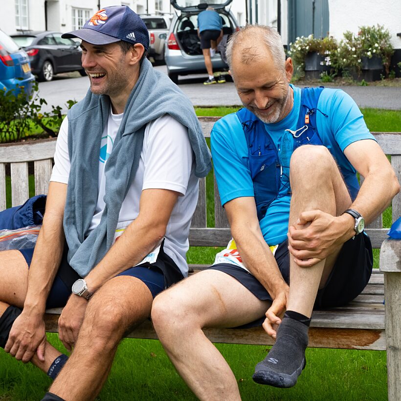 Two male runners sat on a bench changing socks, left one in white top, right in blue top