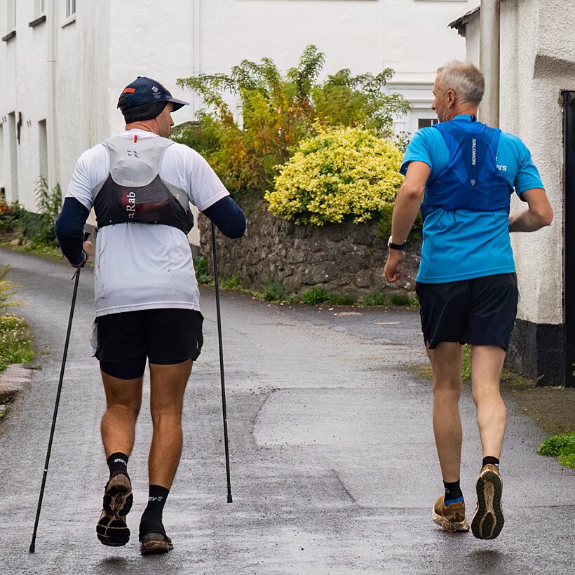 View from the back of two male runners, left one in white top with poles, right in blue top, running up hill