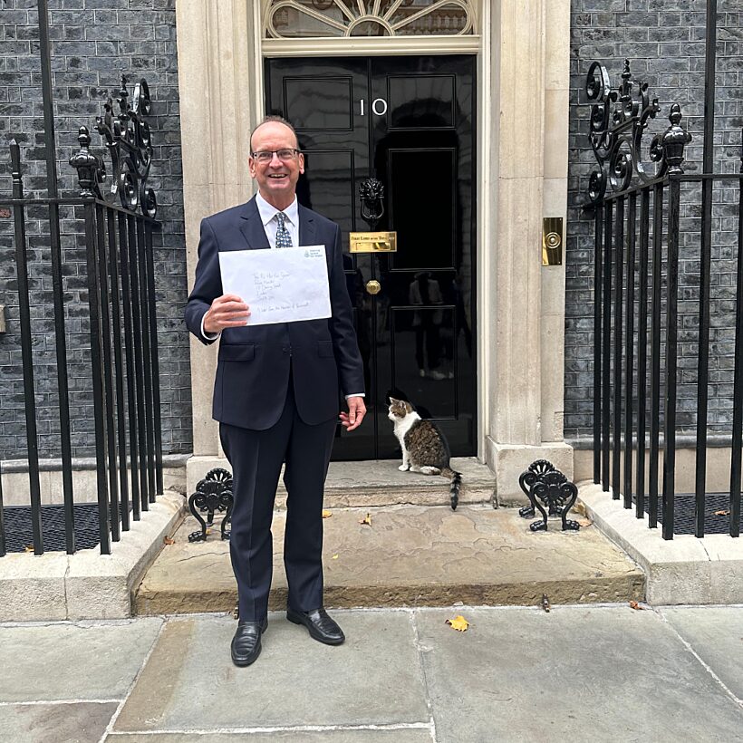 Man in suit holding large envelope in front of black door with cat on the doorstep