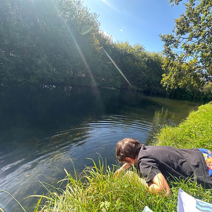 A young boy in a black t-shirt lying on his front with his hand over the bank of a river