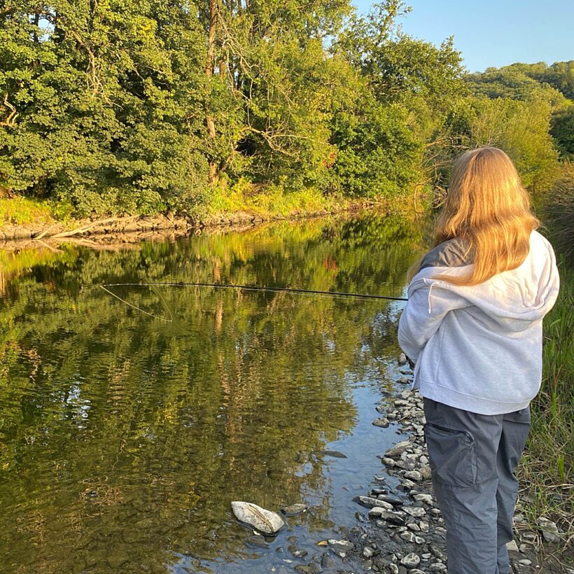 Woman in white hoody stood on gravel river bank with back to camera, fishing