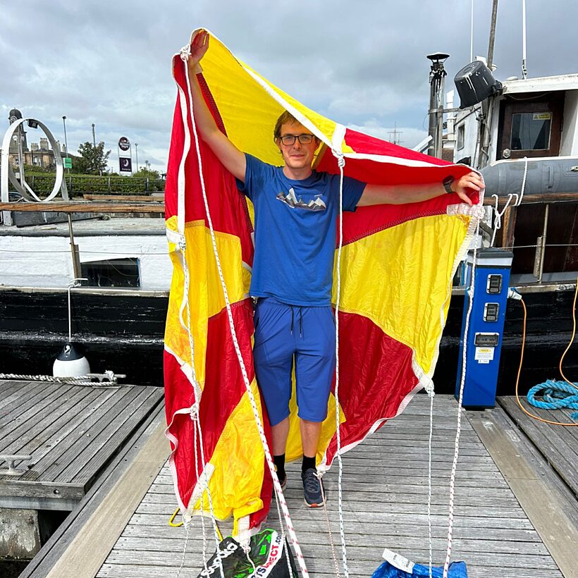 Man in blue top stood on wooden deck holding a yellow and red parachute above his head